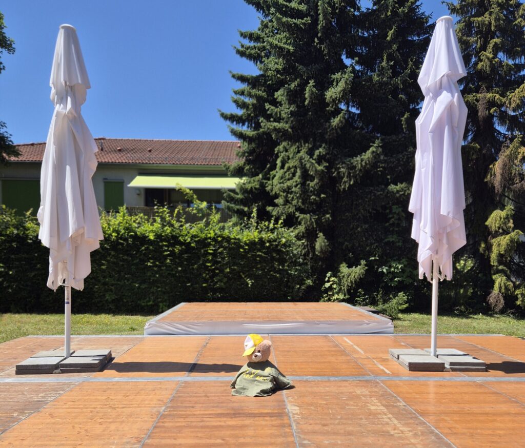Albär, ein Teddybär mit gelber Kappe und grünem T-Shirt, sitzt allein in der Mitte einer leeren Tanzfläche im Freien. Zwei geschlossene weiße Sonnenschirme rahmen ihn ein, im Hintergrund stehen Nadelbäume und eine Hecke unter blauem Himmel.
