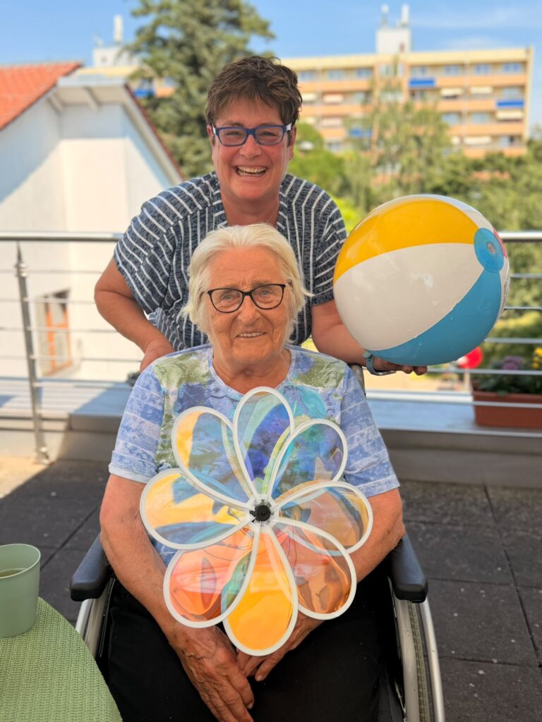 Eine ältere Frau sitzt auf einer Terrasse in einem Rollstuhl und hält ein Windrad in Blumenform. Hinter ihr steht eine weitere Frau und hält einen Wasserball. Beide lachen herzlich.