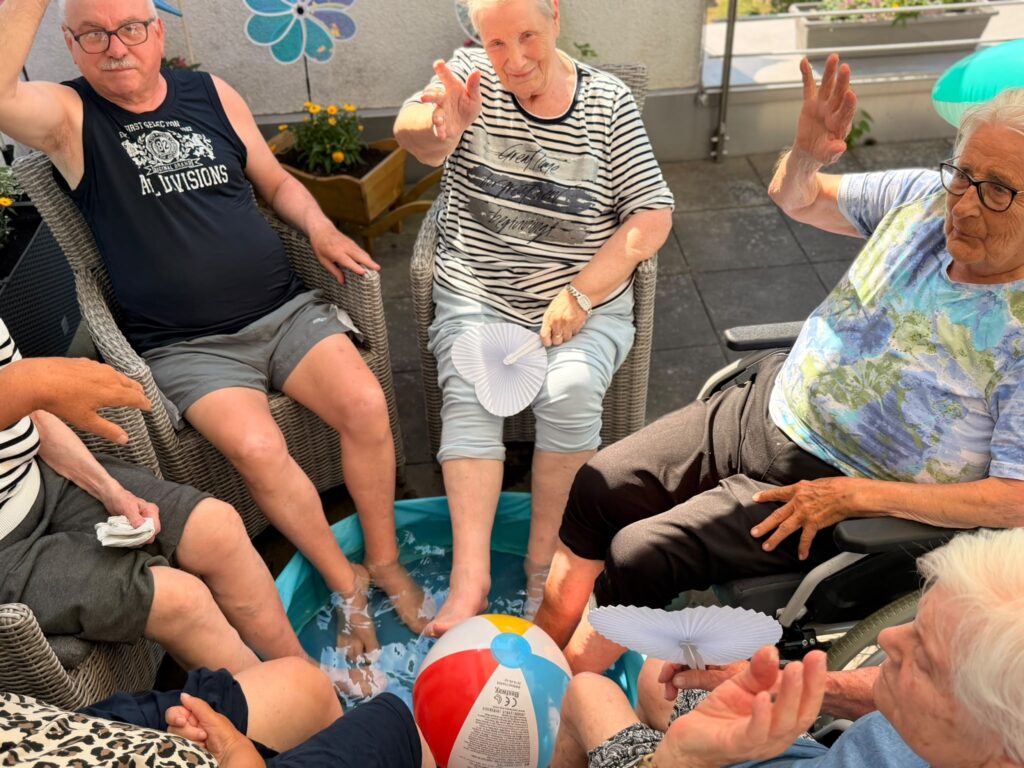 Eine Gruppe älterer Menschen sitzt in einem Stuhlkreis und hält die Füße in ein Planschbecken, das in der Mitte steht.