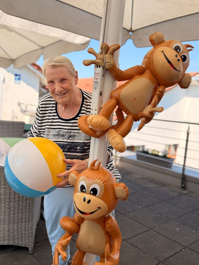 Eine ältere Frau mit weißen Haaren hält einen Wasserball in den Händen. Sie steht unter einem Sonnenschirm, an dem zwei Ballonaffen befestigt sind.