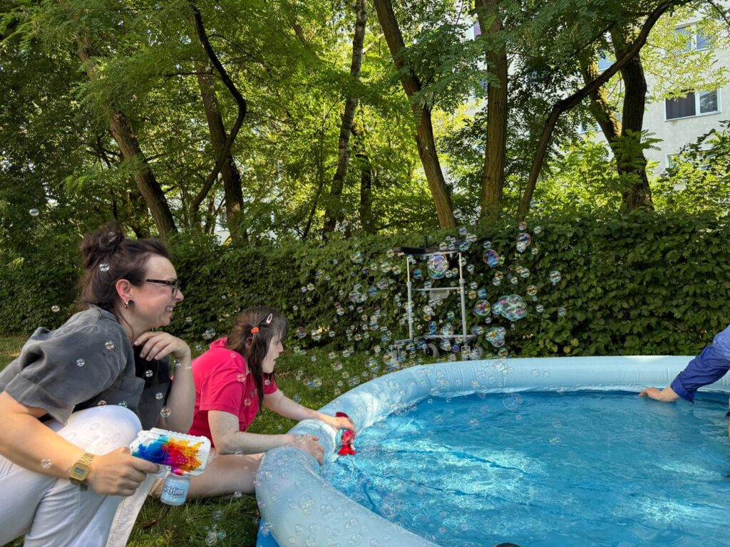 Zwei Frauen hocken vor einem aufblasbaren Pool und spielen mit einer Seifenblasenmaschine.