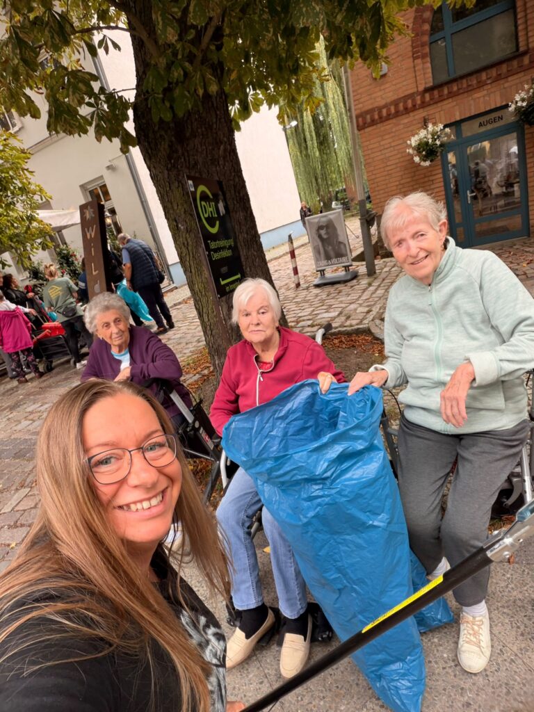 Selfie einer jungen Frau mit zwei älteren Damen im Hintergrund. Sie halten eine Müllgreifzange und einen blauen Müllsack.