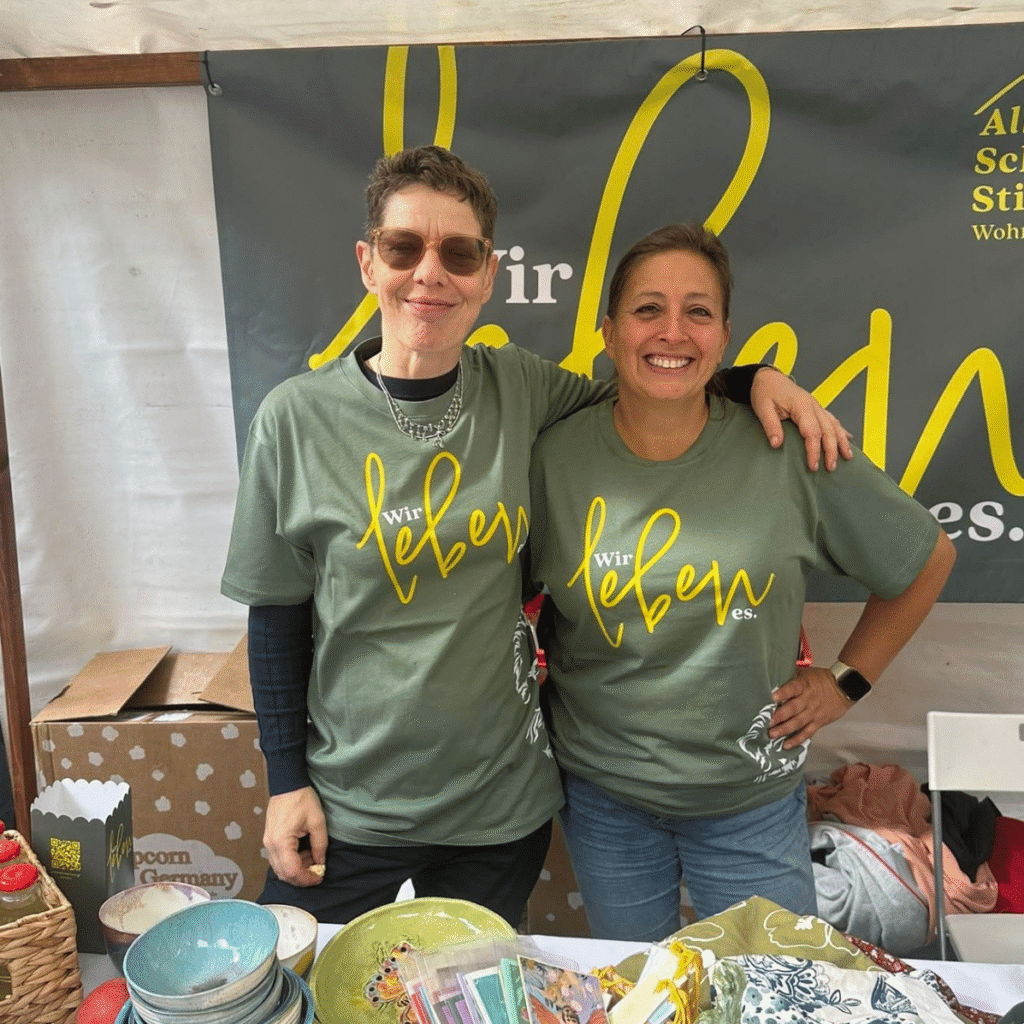 Zwei Frauen stehen Arm in Arm vor einem Banner der Albert Schweitzer Stiftung Wohnen und Betreuen. Sie tragen grüne T-Shirts mit der Aufschrift